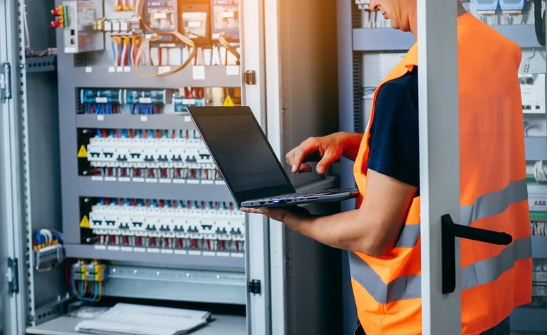 Technician using laptop beside industrial electrical control panel and PLC cabinet